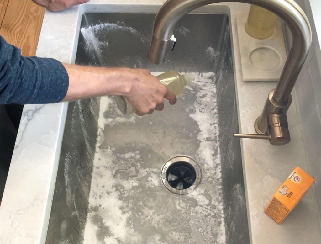 Hand squeezing dish soap into stainless steel sink covered with baking soda